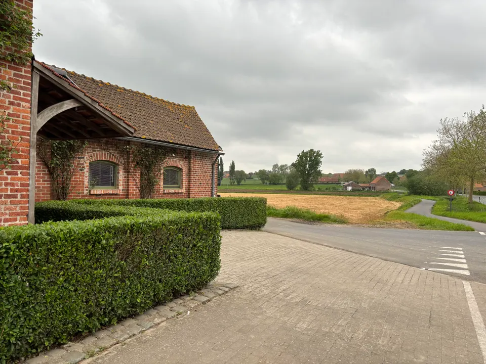 Brick outbuilding with a tiled roof and timber porch behind a hedge, the road and fields beyond