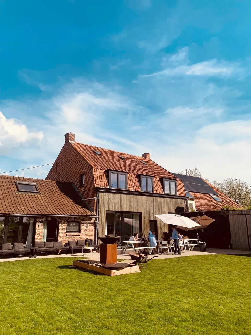 Timber-clad rear facade, terrace with a parasol and guests at the table, and a fire bowl on the lawn