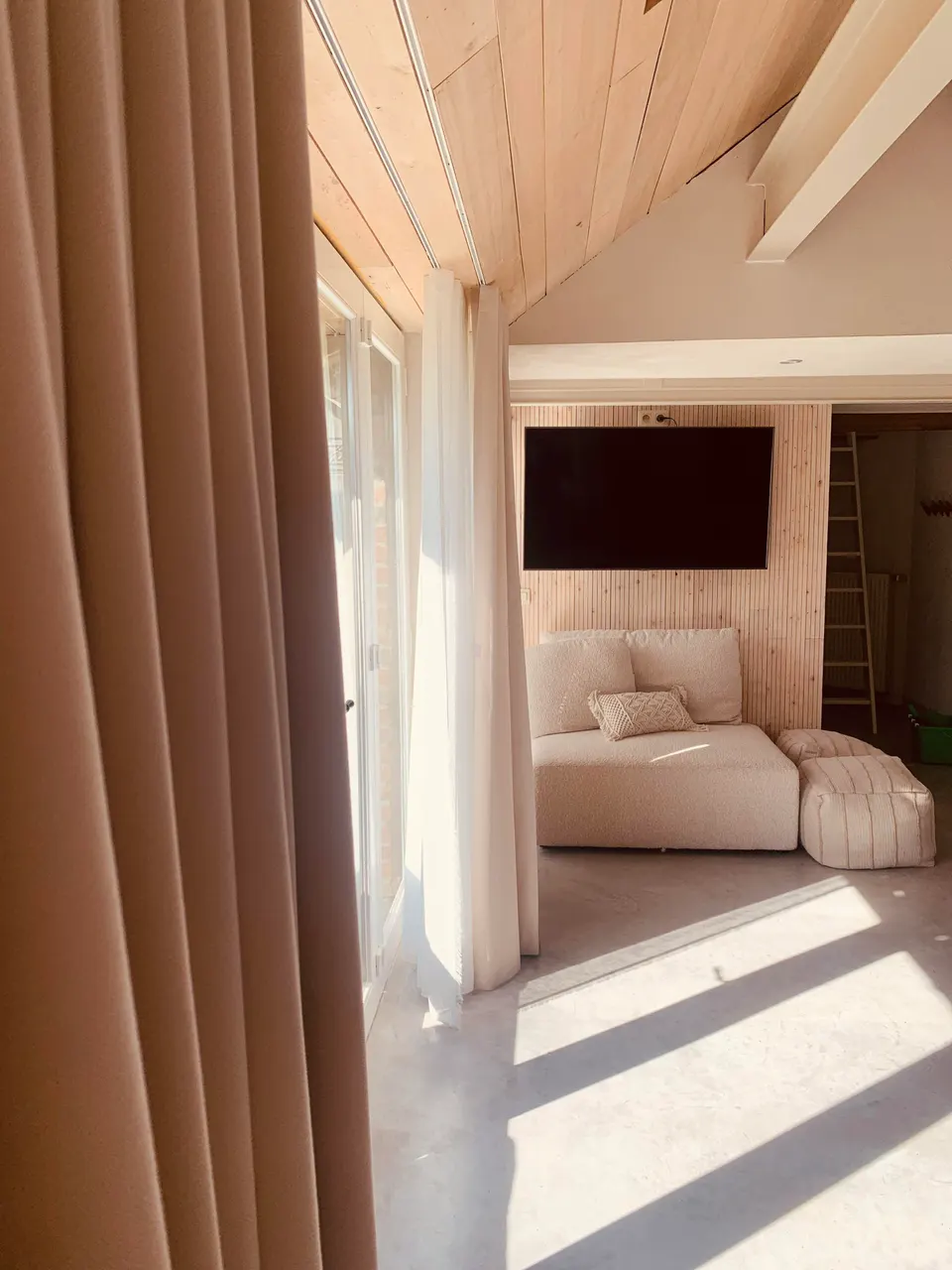 Sunlight on the concrete floor of the living room, with a beige sofa, poufs and a TV on a timber wall
