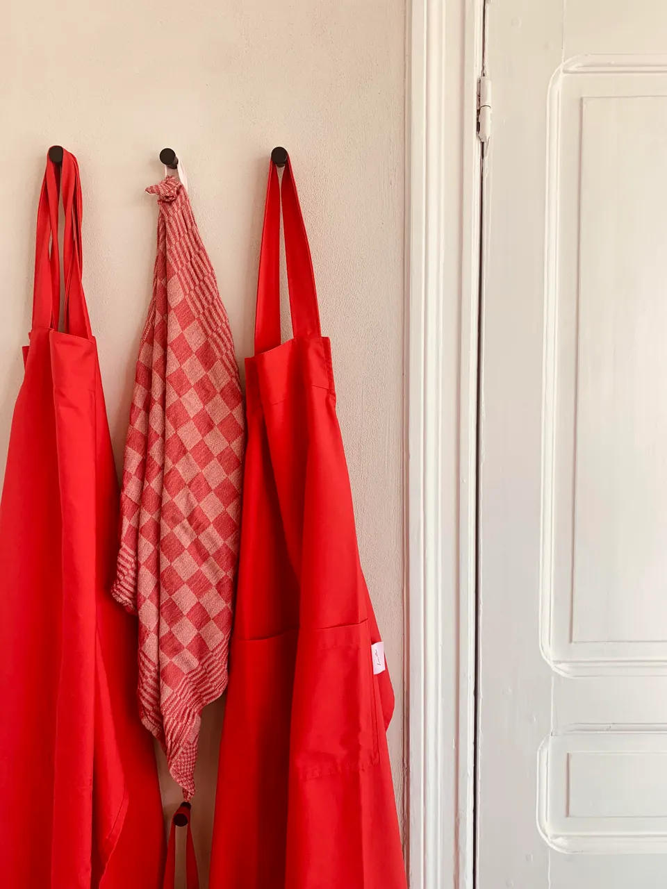 Two red aprons and a red checked tea towel on hooks beside a white door