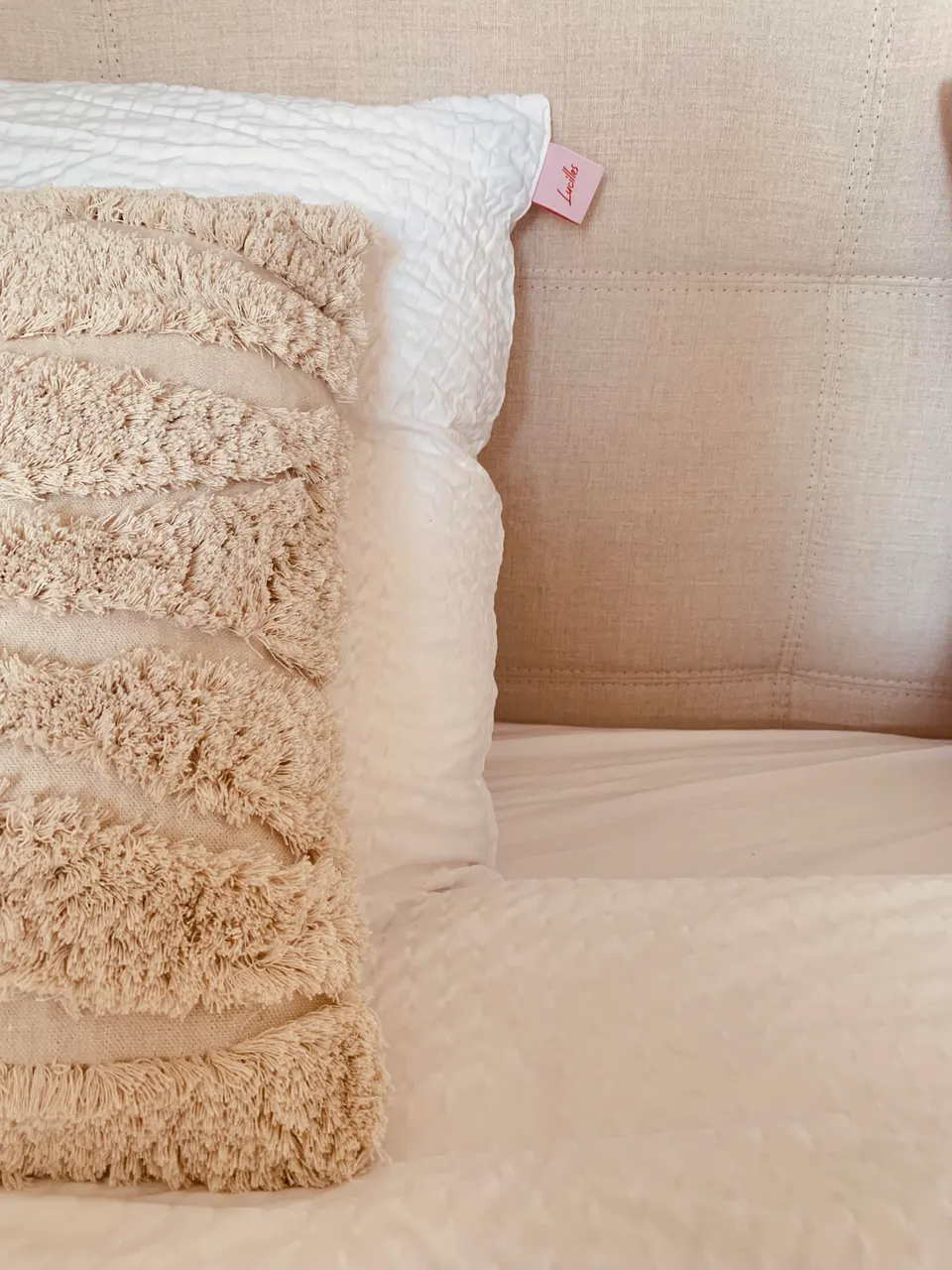 White pillow and a beige tufted cushion on a bed with a linen headboard