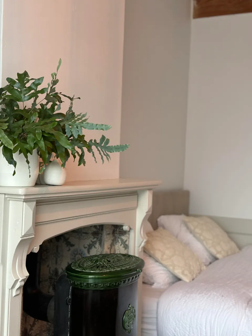 Corner of a bedroom with a white mantelpiece, a green stove, a plant and a made bed