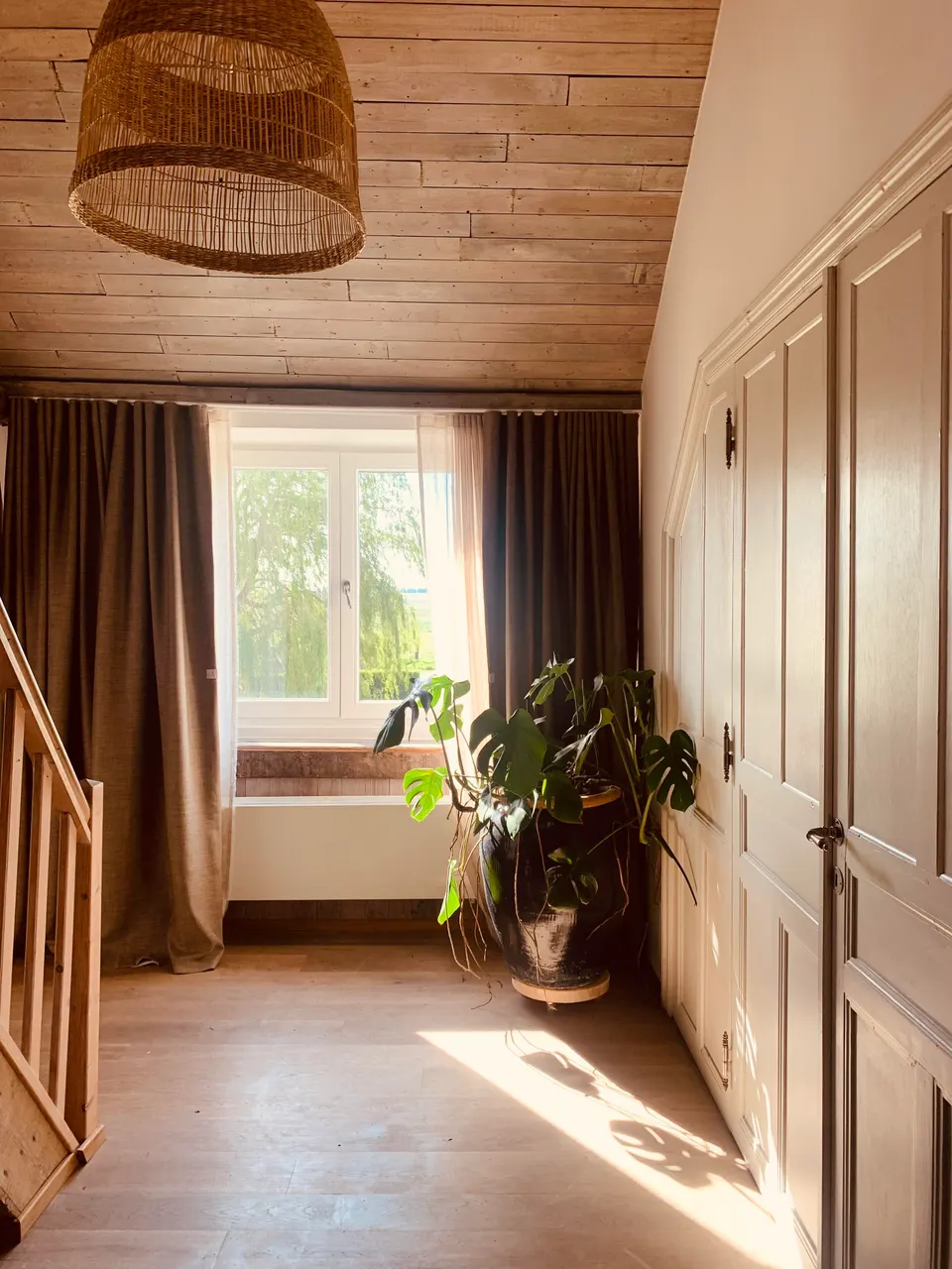 Landing under the roof with a wooden ceiling, rattan lamp, built-in cupboards, a plant and a window