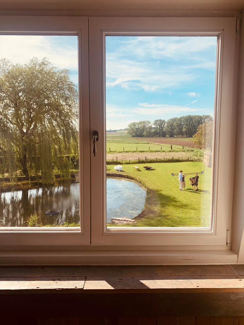 View through a window of the garden: pond, weeping willow, people playing on the lawn, meadows beyond