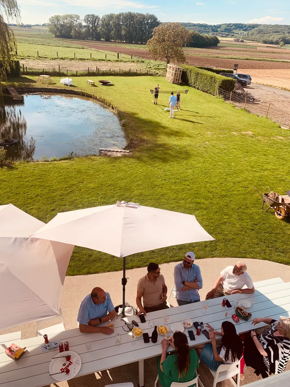 A group of guests at a long white table under parasols, with the pond, lawn and fields behind
