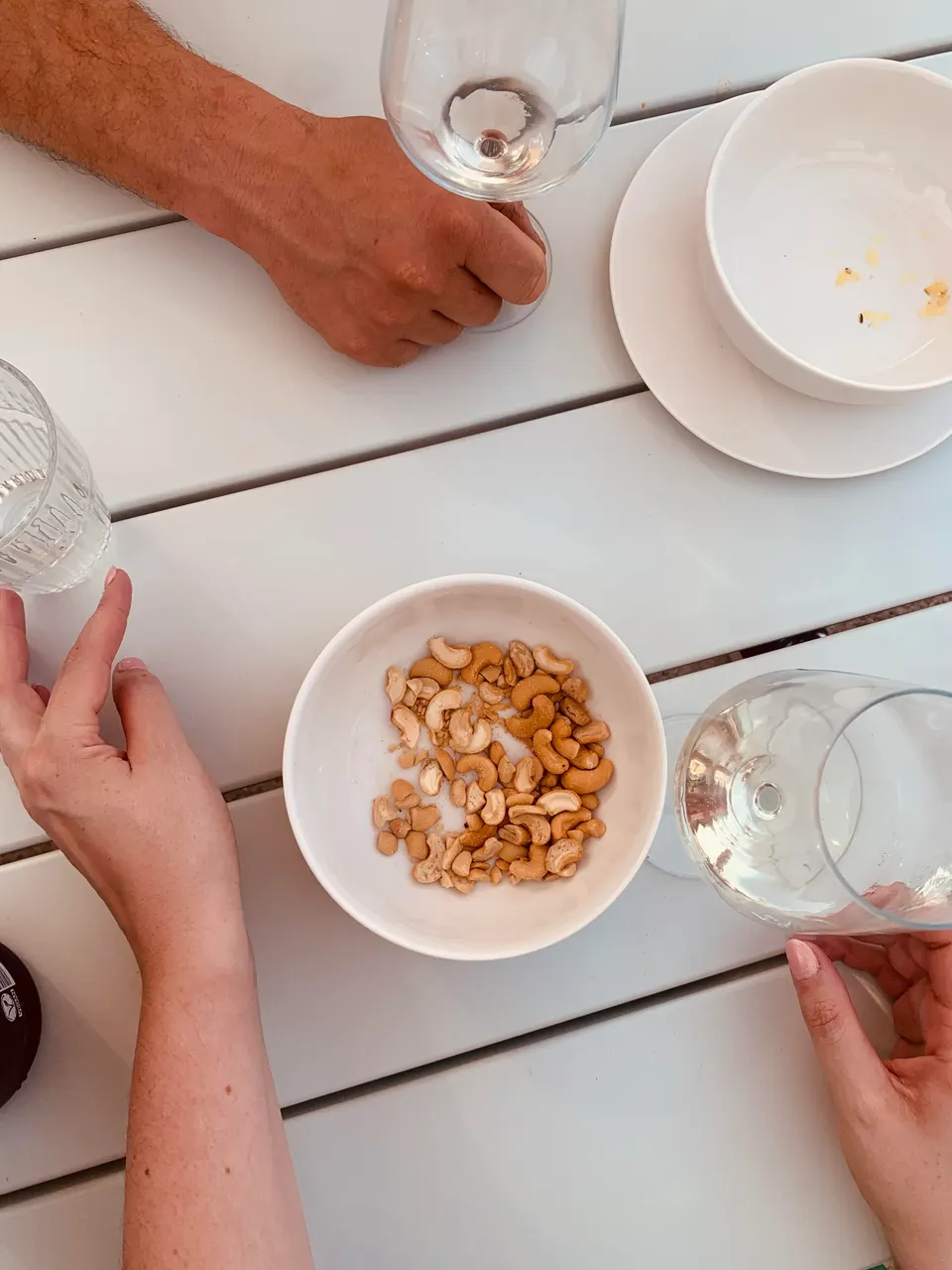 Hands around wine glasses and a bowl of cashew nuts on a white wooden table