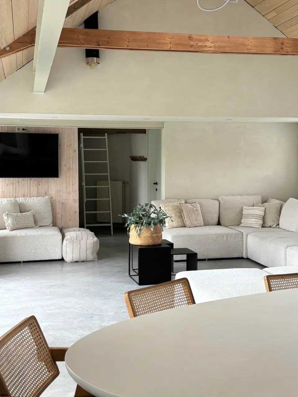 Seating area with beige sofas and a wall-mounted TV, a round dining table and cane chairs in front