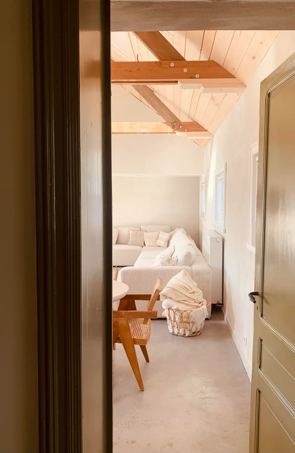 View through an open door into a bright sitting area with a white sofa and timber beams