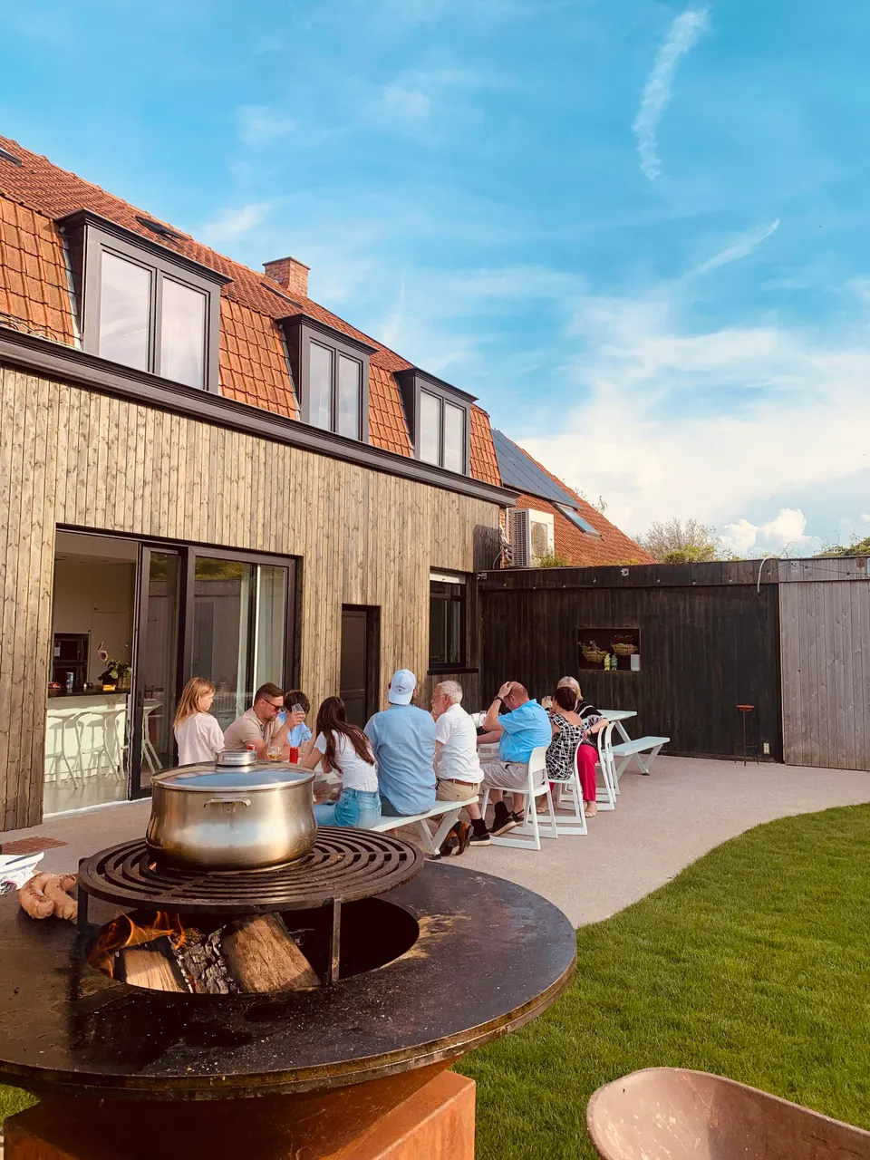 A cooking pot on a wood-fired fire bowl, with guests at a long table on the terrace behind