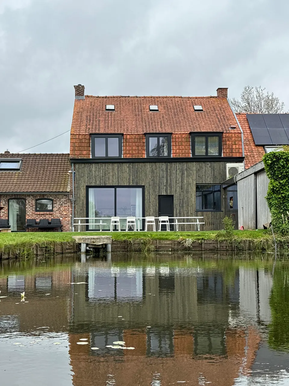 Rear of the house with timber cladding and a terrace with white tables, reflected in the pond