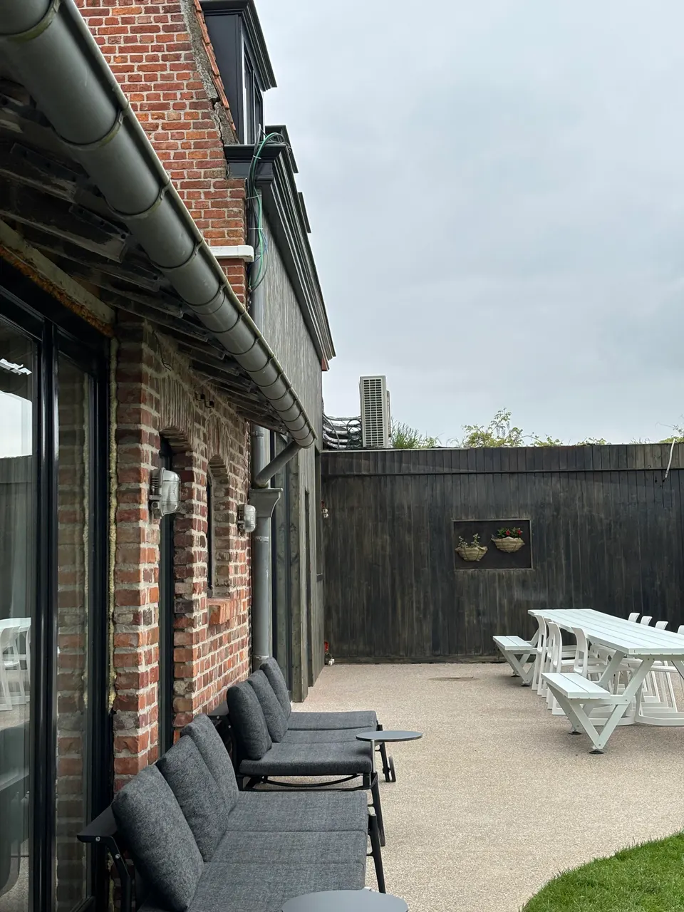 Terrace with grey lounge sofas against the wall and white picnic tables by a dark timber fence
