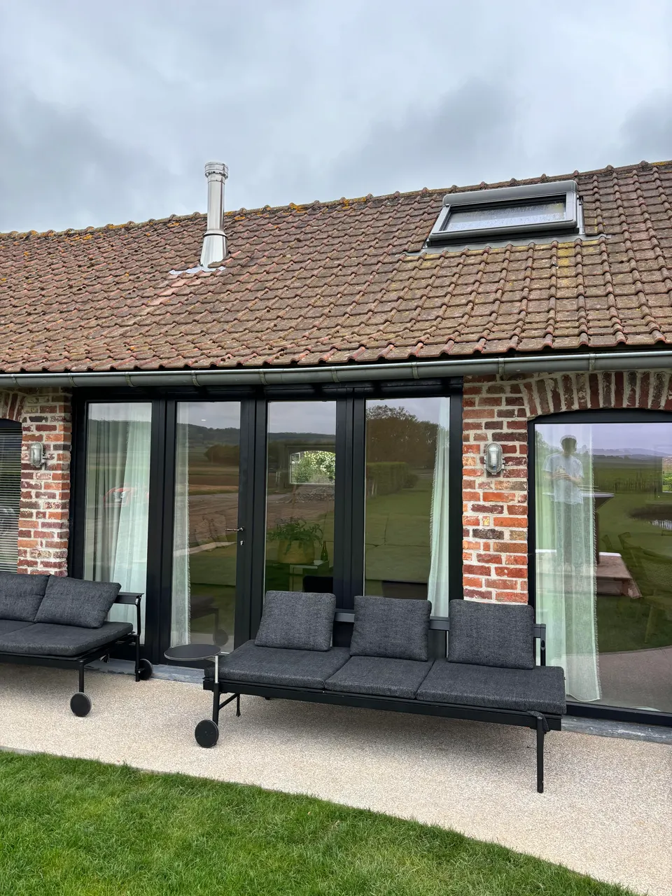 Brick rear wall with large glass sliding doors, grey garden sofas on the terrace and a strip of lawn