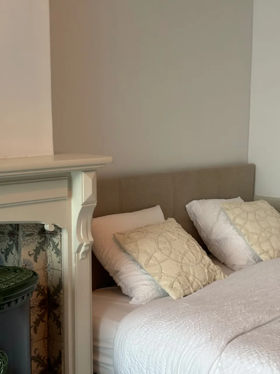 Bedroom with a double bed, white bedspread and cushions beside a white mantelpiece with a green stove