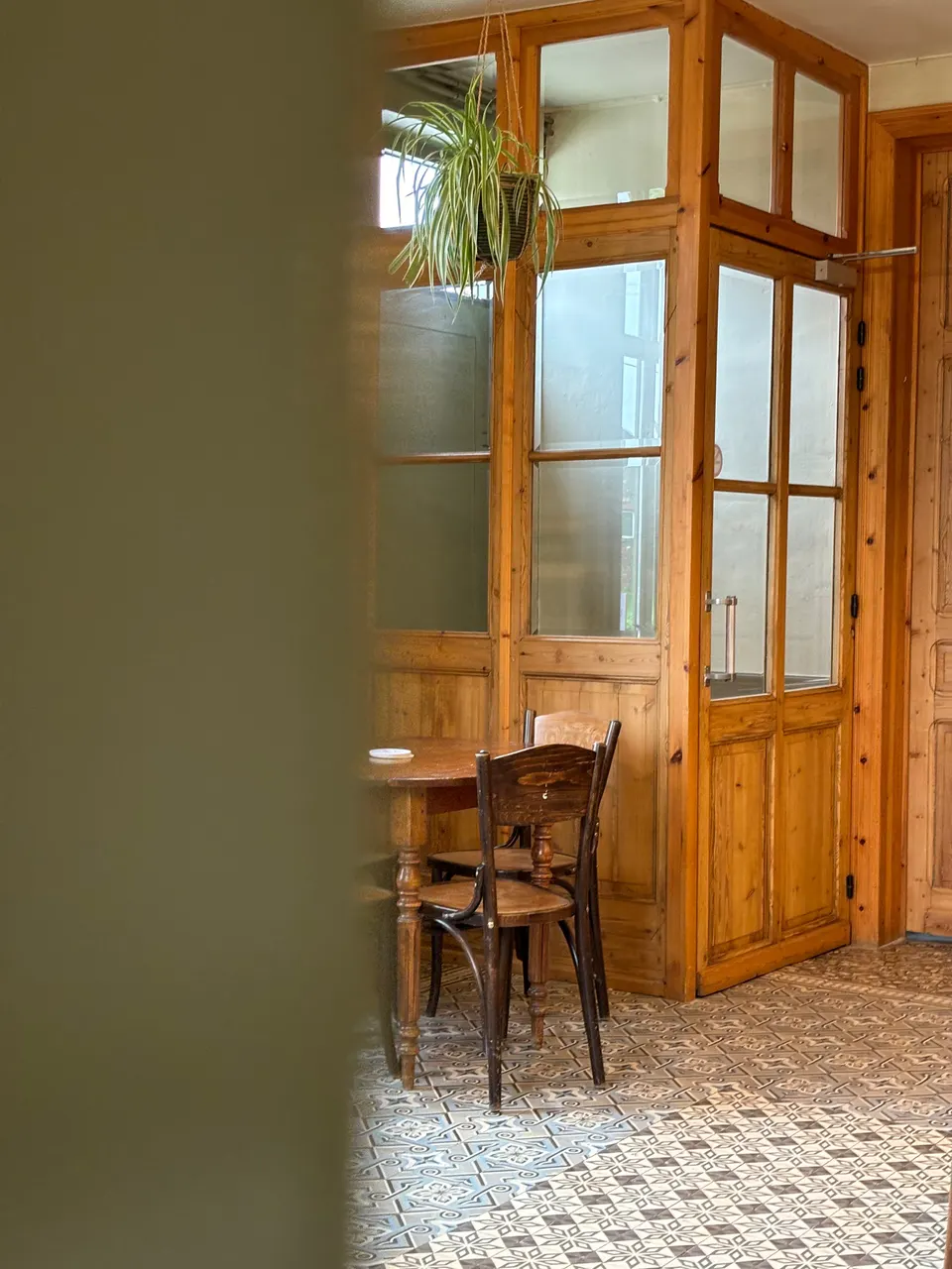 Corner of the café with wooden glazed doors, a hanging plant, a small table and chairs and a cement-tile floor