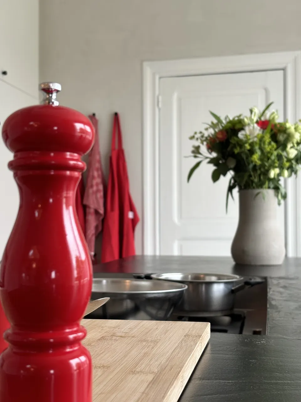 Kitchen with a red pepper mill, chopping board, pans on the hob, a vase of flowers and red aprons