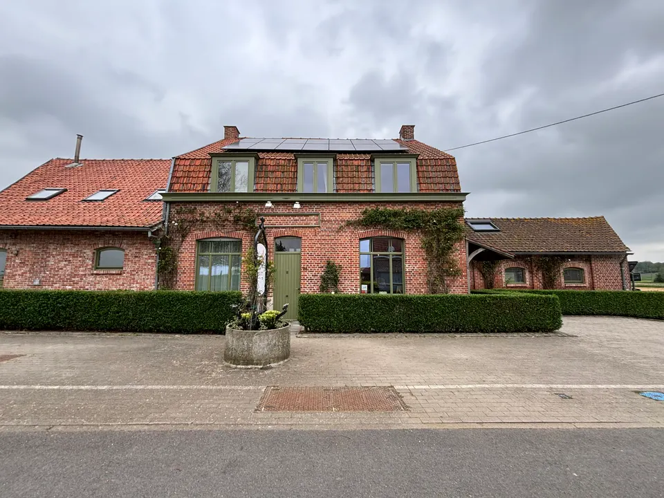 Façade de Lucilles : brique rouge, fenêtres et porte vertes, lucarnes, haies et parvis pavé