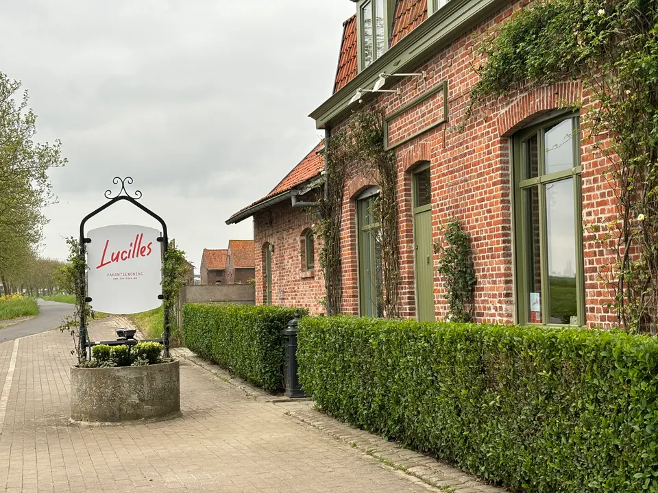Façade en brique avec plantes grimpantes et haies taillées le long de la rue, et l’enseigne en fer forgé de Lucilles