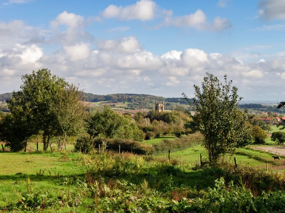 Rolling green landscape with trees and meadows, a church tower in the distance under a cloudy sky